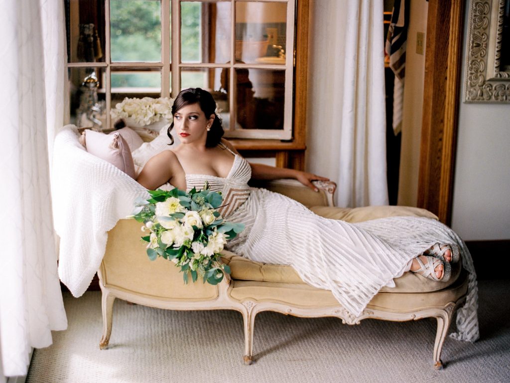The bride lounges on a couch with her bouquet near a window.
