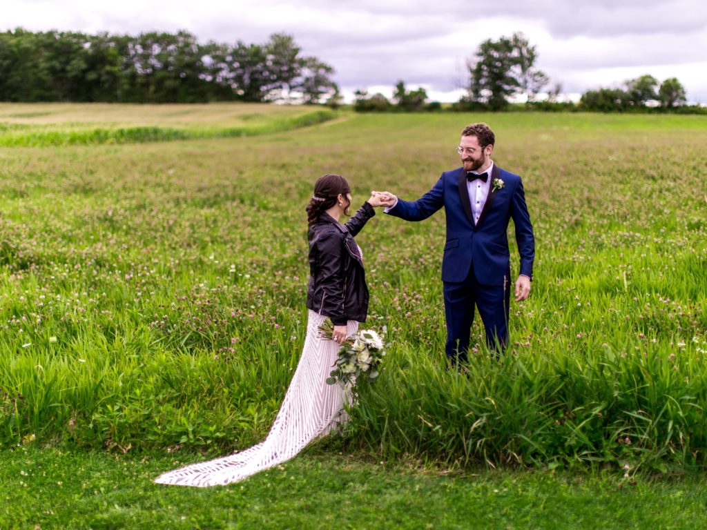 The bride and groom walk through a field. The bride wears a leather jacket over her dress.