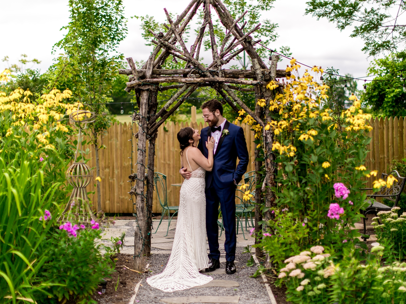 The bride and groom stand in a garden full of flowers. They stand beneath a wood arbor.