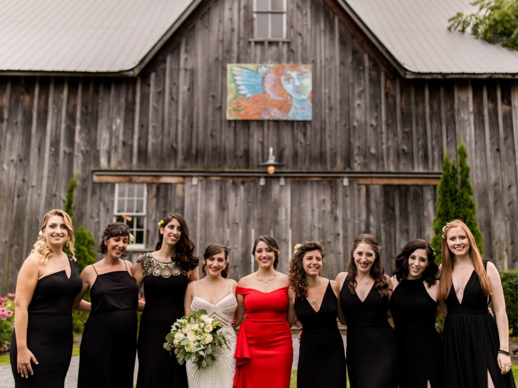 The bride stands in front of the barn with her bridesmaids. 