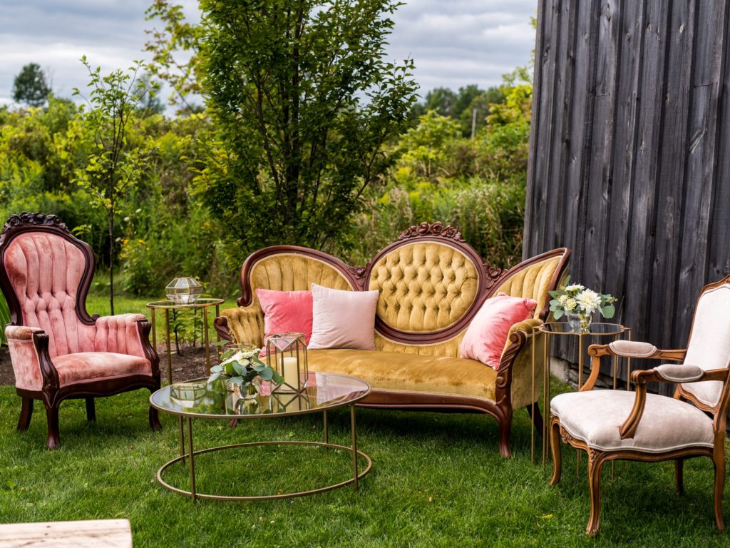 Vintage furniture in the grass, including a pink chair, yellow couch, and white chair with a glass coffee table