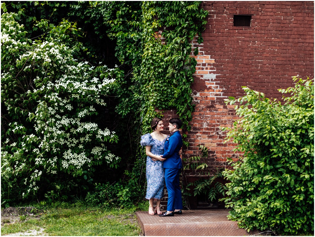 A couple poses in front of a brick wall beside white flowers.